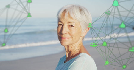Image of network of connections with icons over smiling senior woman on beach. digital interface,...