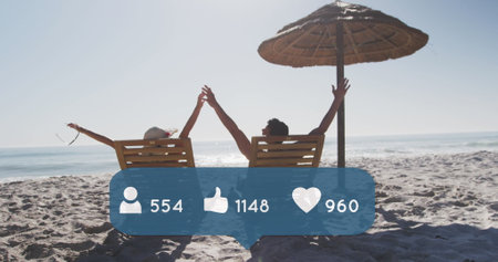 Image of people, thumbs up and heart icons and numbers over couple in deckchairs on beach. digital interface, social media and global technology concept digitally generated image.の写真素材