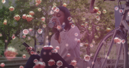 Multiple covid-19 cells floating against woman with lowered face mask drinking coffee in the park....