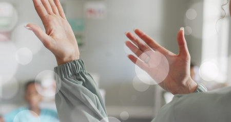 Image of white bokeh spots over hands of diverse female teacher and pupils in class. Communication, teaching, school, education, childhood and learning, digitally generated image.の写真素材