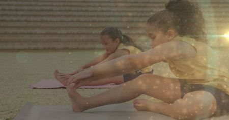 Image of bokeh lights over diverse schoolgirls exercising in outdoor yoga class. Health, school, education, childhood and learning, digitally generated image.の写真素材