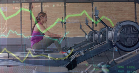 Image of data on graph over biracial woman training on rowing machine at gym. Fitness, exercise, strength, data, digital interface and technology digitally generated image.の写真素材