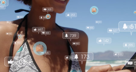 Social media icons floating against african american woman using smartphone at the beach. social media networking and technology conceptの写真素材