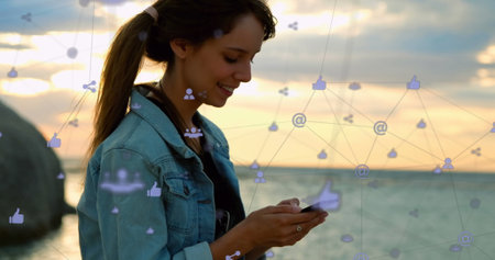 Network of digital icons against caucasian woman using smartphone sitting near the sea. global...