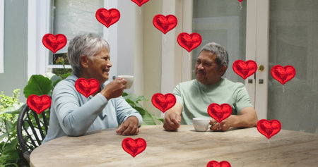 Image of red hearts over senior couple drinking coffee at home. digital interface, social media,...