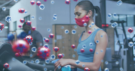 Covid-19 cells floating against woman wearing face mask cleaning gym equipment with disinfectant. coronavirus covid-19 pandemic and fitness conceptの写真素材