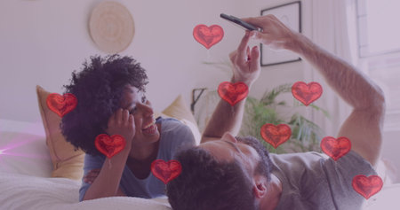 Image of hearts over diverse couple using smartphone in bed. Relationship, love, romance and lifestyle concept digitally generated image.の写真素材