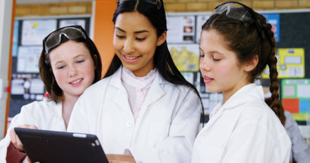 Three teenage girls are engaged with a tablet in a classroom setting. Dressed in white lab coats, the biracial girl with dark hair smiles as she shows something to her Caucasian peers with light brown and red hair.の写真素材