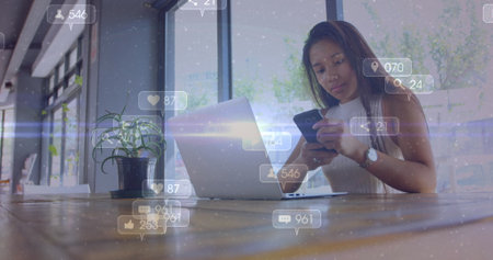 Image of social media icons with numbers over biracial woman using smartphone. Global social media, technology and digital interface concept digitally generated image.の写真素材