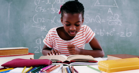 Image of mathematical equations floating over schoolchild sitting at desk and reading a book....