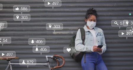 Image of social media icons and numbers on banners over woman in face mask using smartphone. global...