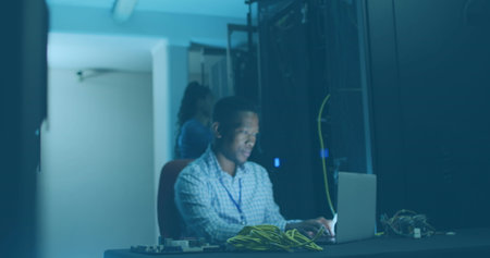 African American father wearing blue shirt, holding cables, working on a laptop. Smiling, focusing...