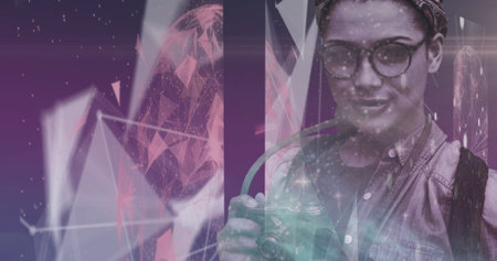 Biracial young woman holding camera, standing against abstract background. Wearing glasses and denim vest, her hair tied back, capturing momentsの写真素材