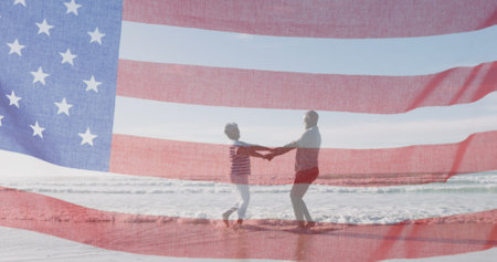 Diverse couple holding hands on beach, American flag overlaying. Both seniors, wearing casual summer clothes, enjoying sunny dayの写真素材