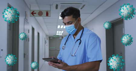 Hispanic male doctor in blue scrubs checks a tablet in a hospital corridor. He has dark hair and a...