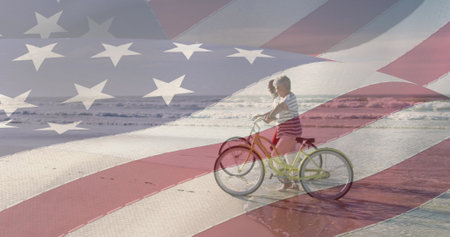A young Caucasian boy is riding bicycle on a beach, overlaid with American flag. He wears a striped shirt and shorts, enjoying a sunny day outdoorsの写真素材