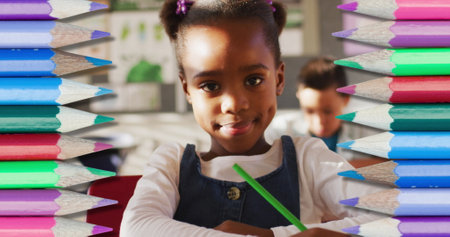 African American girl with green pencil, Caucasian boy behind in school. She has dark hair in pigtails, wearing blue overalls; he focuses on writingの写真素材