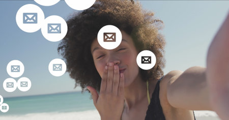 Image of envelope icons over biracial woman holding camera and blowing kiss at beach. Digital...