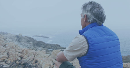 Caucasian man gazes at the sea from a rocky shore, with copy space. His contemplative stance...