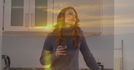 Image of sunset landscape over caucasian woman drinking coffee in kitchen. Lifestyle and domestic...