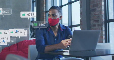 Image of social media icons and numbers over biracial businessman in face mask with laptop. Global...