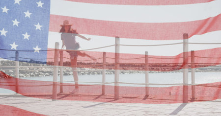 American flag waving against caucasian woman wearing a hat climbing over the wooden pier fence. american independence and celebration conceptの写真素材