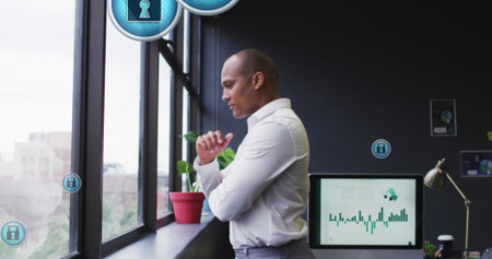 Image of security padlock icons floating against biracial man looking out of window at office. Cyber...