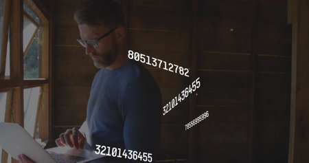 Image of multiple changing numbers over caucasian man standing near window and using laptop. Digital composite, multiple exposure, business, growth, digits and technology concept.の写真素材