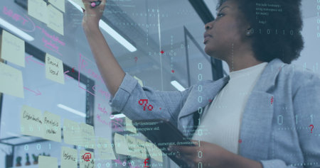 Image of computer language, binary codes over african american woman sticking note on glass wall....