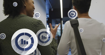 Image of multiple bar graph icons floating against two diverse women discussing at office. Business...