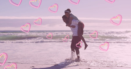 Image of pink hearts over happy african american couple piggybacking on beach. Love, togetherness, romance and holidays concept digitally generated image.の写真素材