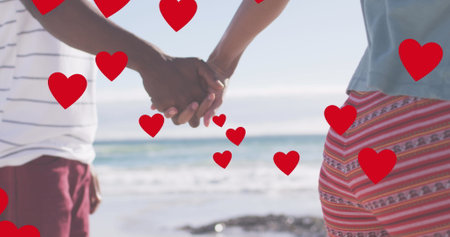 Image of red hearts over midsection of african american couple holding hands on beach. Love, togetherness, romance and holidays concept digitally generated image.の写真素材
