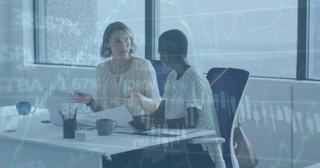 Image of graphs and trading board over diverse female coworkers discussing reports on laptop. Digital composite, multiple exposure, business, planning, teamwork, stock market and technology.の写真素材