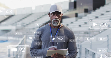 Image of notification icons with changing numbers over african american coach using tablet. Digital composite, sports, competition, stadium, training, mobile apps, social media, technology.の写真素材