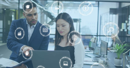 Image of icon in circles over diverse coworkers standing and discussing reports on laptop. Digital composite, multiple exposure, globalization, education, planning, teamwork and technology.の写真素材