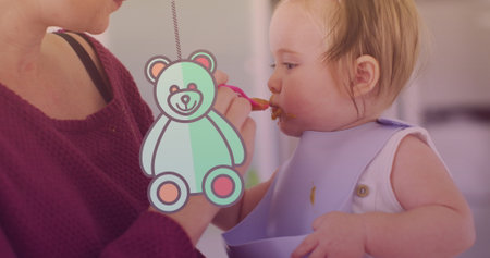 Image of teddy bear icon over caucasian mother feeding her baby with spoon. national weaning week and parenthood concept digitally generated image.の写真素材