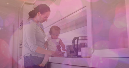 Image of pink spots of light over caucasian woman with baby in kitchen. national weaning week and parenthood concept digitally generated image.の写真素材