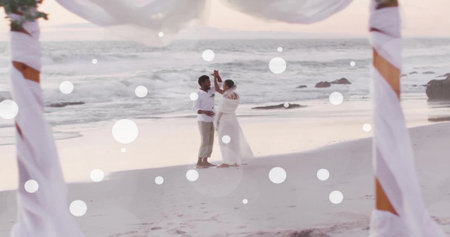 Image of white spots over african american bride and groom dancing on beach at wedding. Romance, love, vacations and marriage, digitally generated image.の写真素材