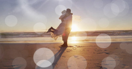 Image of light spots over happy african american groom lifting bride on sunset beach at wedding. Romance, love, vacations and marriage, digitally generated image.の写真素材