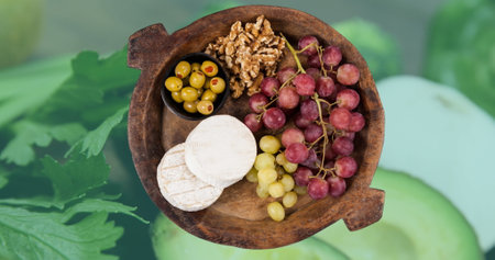 Image of wooden bowl of cheese, grapes, nuts and olives over green vegetables. World food day concept digitally generated image.の写真素材