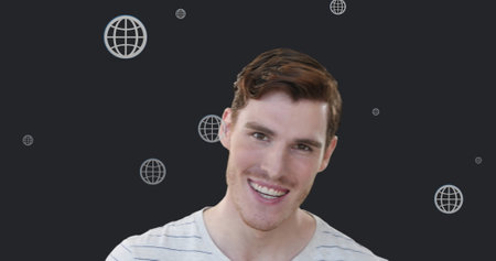 Image of multiple web globe icons floating over caucasian man smiling against grey background. Global networking and business technology conceptの写真素材