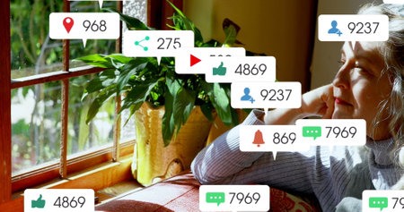 Image of social media icons over caucasian senior woman looking out of window at home. Social media networking technology conceptの写真素材