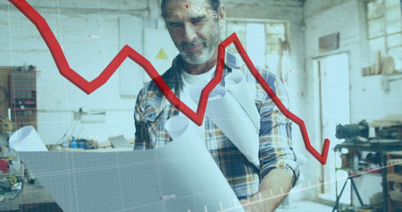 Image of red graph processing over mature caucasian male carpenter with blueprints in workshop. Connection, network, data, small business and carpentry, digitally generated image.の写真素材