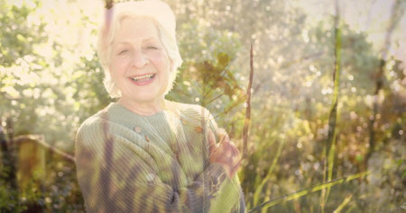 Image of spots of light and trees over smiling senior caucasian woman in garden. Happiness, senior lifestyle, active lifestyle and retirement concept digitally generated image.の写真素材