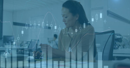 Asian woman, middle-aged, wearing light blue shirt, analyzing data on computer. Focused on screen,...