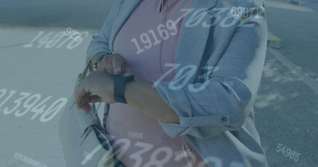 Image of multiple changing numbers over biracial woman standing and using smartwatch. Digital composite, multiple exposure, business, growth, abstract and technology concept.の写真素材