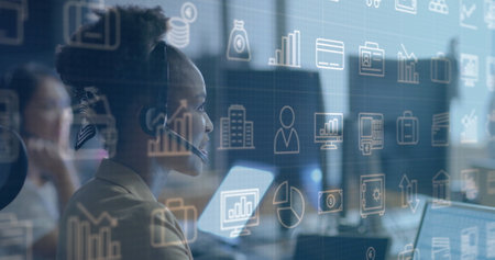 Image of business icons over african american businesswoman using headset and laptop at office. Work, connection, business, digital interface and communication, digitally generated image.の写真素材