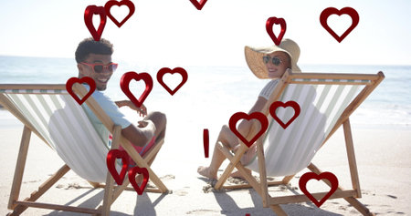 Image of hearts moving over diverse couple in love in deckchairs on beach in summer. Love, romance, valentine's day and vacation concept digitally generated image.の写真素材