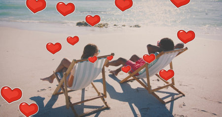Image of hearts moving over diverse couple in love in deckchairs on beach in summer. Love, romance, valentine&#39;s day and vacation concept digitally generated image.の写真素材