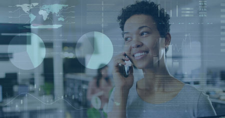 Image of infographic interface over smiling biracial woman talking on cellphone in office. Digital composite, multiple exposure, report, business, growth, progress and technology concept.の写真素材